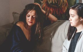 two young women sit on a brown couch. The one on the left has brown hair and is wearing a sparkly blue shirt and a flower patterned skirt. The one of the right had black hair and is wearing a ivory sweater over a charcoal grey top