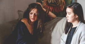 two young women sit on a brown couch. The one on the left has brown hair and is wearing a sparkly blue shirt and a flower patterned skirt. The one of the right had black hair and is wearing a ivory sweater over a charcoal grey top