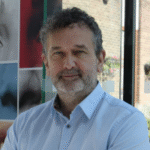 a middle-aged white man with short curly dark hair and a slight silver beard wearing a light blue button-up shirt