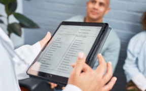 Photo of a tablet in foreground being used to check in patients seated in image background.