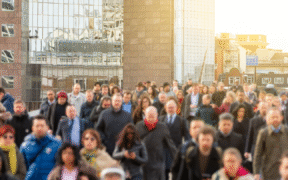 Photo of lots of people crossing bridge with faces blurred out. Bright reflection on buildings in background.