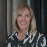 a white woman with shoulder-length blond hair, wearing a button-up shirt that has a pattern of black and white splotches