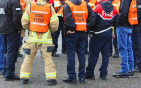 a group of people cropped from the shoulders down with jackets that say “Fire” and other disaster-response-related things on them, standing on gravel