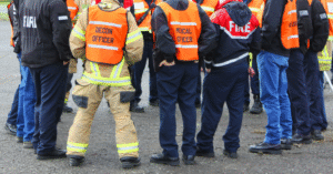 a group of people cropped from the shoulders down with jackets that say “Fire” and other disaster-response-related things on them, standing on gravel