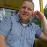 A white middle-aged man with cropped blond hair wearing a short-sleeve light blue button up shirt, sitting and leaning on one elbow