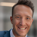 a white man with a tuft of brownish-black hair, wearing a blue suit