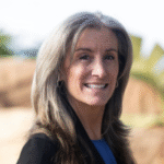an older woman with shoulder-length brownish gray hair, wearing a blue top with a black jacket over it