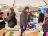eight people at event distributing Narcan and other supplies, brown paper bags and tubs of supplies in foreground and people in coats interacting under a gray tent