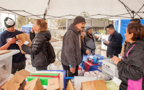 eight people at event distributing Narcan and other supplies, brown paper bags and tubs of supplies in foreground and people in coats interacting under a gray tent
