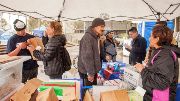 eight people at event distributing Narcan and other supplies, brown paper bags and tubs of supplies in foreground and people in coats interacting under a gray tent