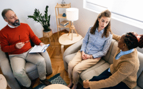 a photo with seated therapist on the left in red sweater talking to two women on couch on the right side of image, who are looking at and talking to each other
