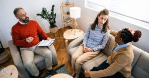 a photo with seated therapist on the left in red sweater talking to two women on couch on the right side of image, who are looking at and talking to each other