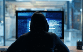 photo of the back of person in black hood looking at hacker-type computer data on screen
