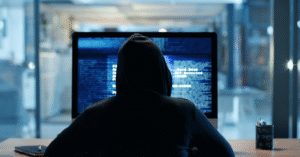 photo of the back of person in black hood looking at hacker-type computer data on screen