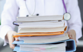 a photo of person in a white lab coat with stethoscope holding a big pile of binders and books