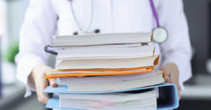 a photo of person in a white lab coat with stethoscope holding a big pile of binders and books