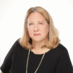 a white woman with shoulder-length light blond hair wearing a black top and necklace